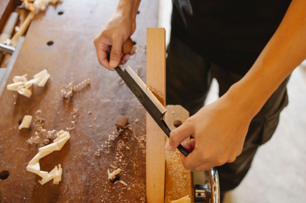 Falegname al banco da lavoro mentre pialla un asse di legno, mani e utensile in primo piano.